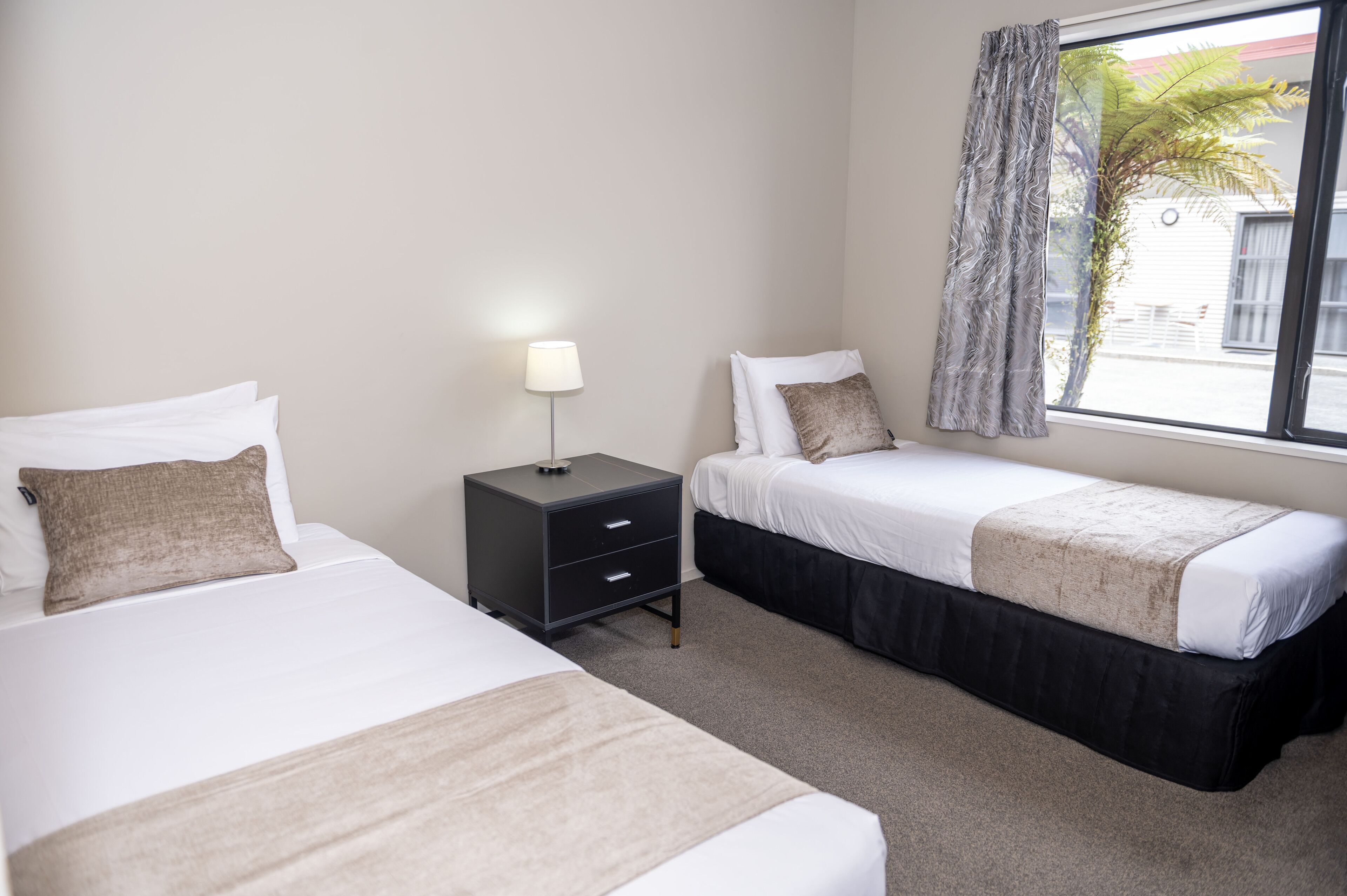 A hotel room with two beds, a nightstand, a lamp, and a window with curtains.