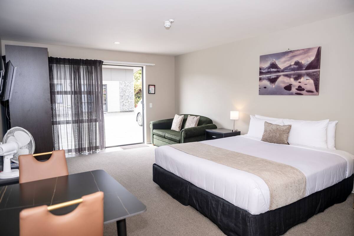 A hotel room with a bed, a desk, a chair, a fan, a green sofa, and a painting on the wall.