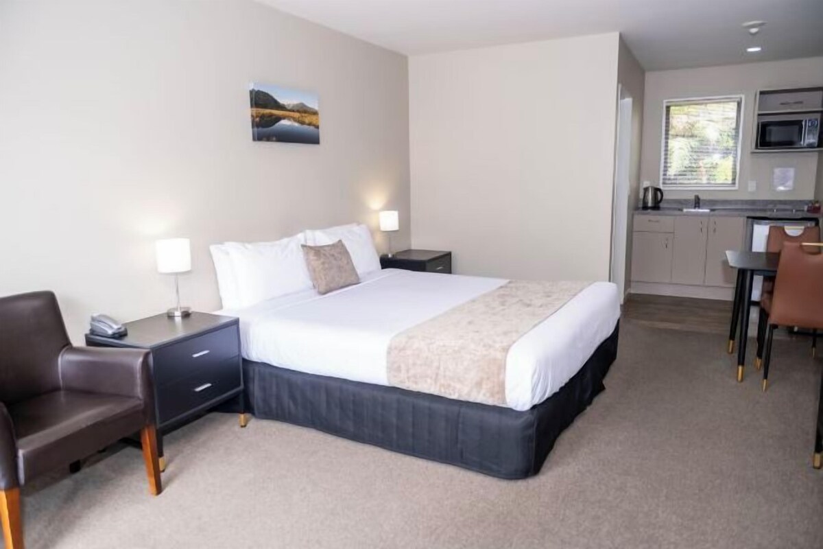 A hotel room with a large bed, a desk, a chair, a nightstand, a lamp, a picture on the wall, and a kitchenette with a microwave and sink.