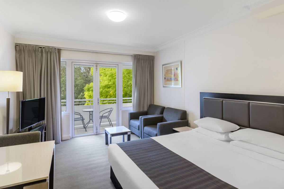 A hotel room with a bed, a desk, a chair, a TV, and a balcony with a view of trees.