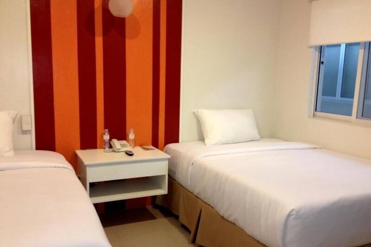 A hotel room with two beds, a small bedside table with a phone and a bottle, and a window with blinds.