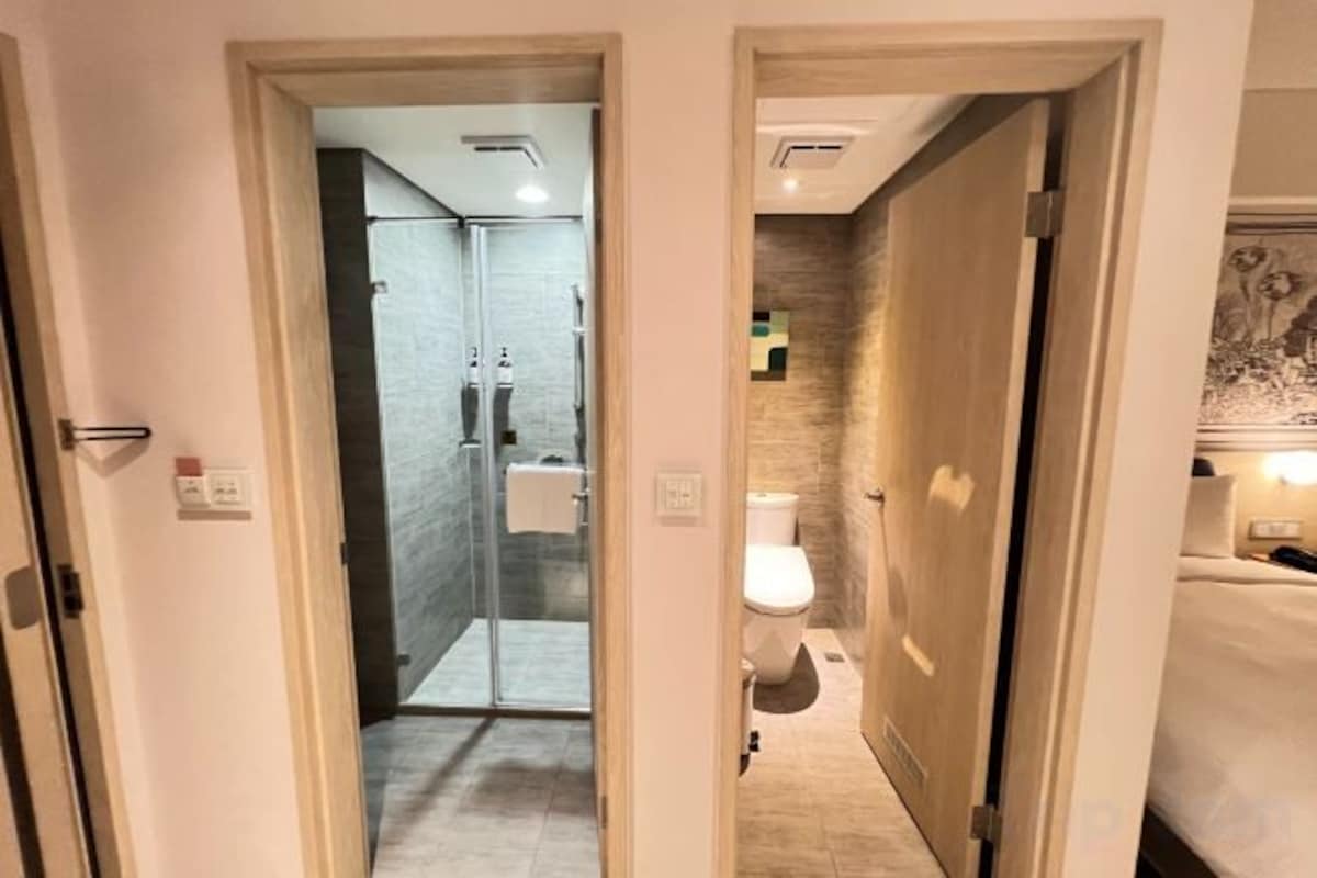 A hotel bathroom with a shower, toilet, and a bed visible through an open door.