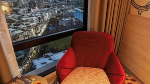 A modern room with a red velvet armchair and ottoman, a small round table with a book and a cup, and a large window offering a city view.