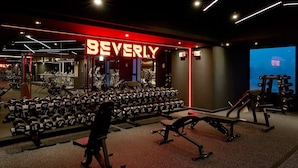 A gym with a 'BEVERLY' sign, weightlifting equipment, treadmills, and a digital display.