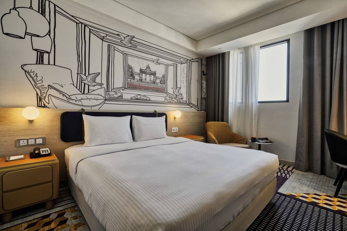 A hotel room with a large bed, a nightstand with a lamp, a chair, a small table with a cup, and a wall mural.