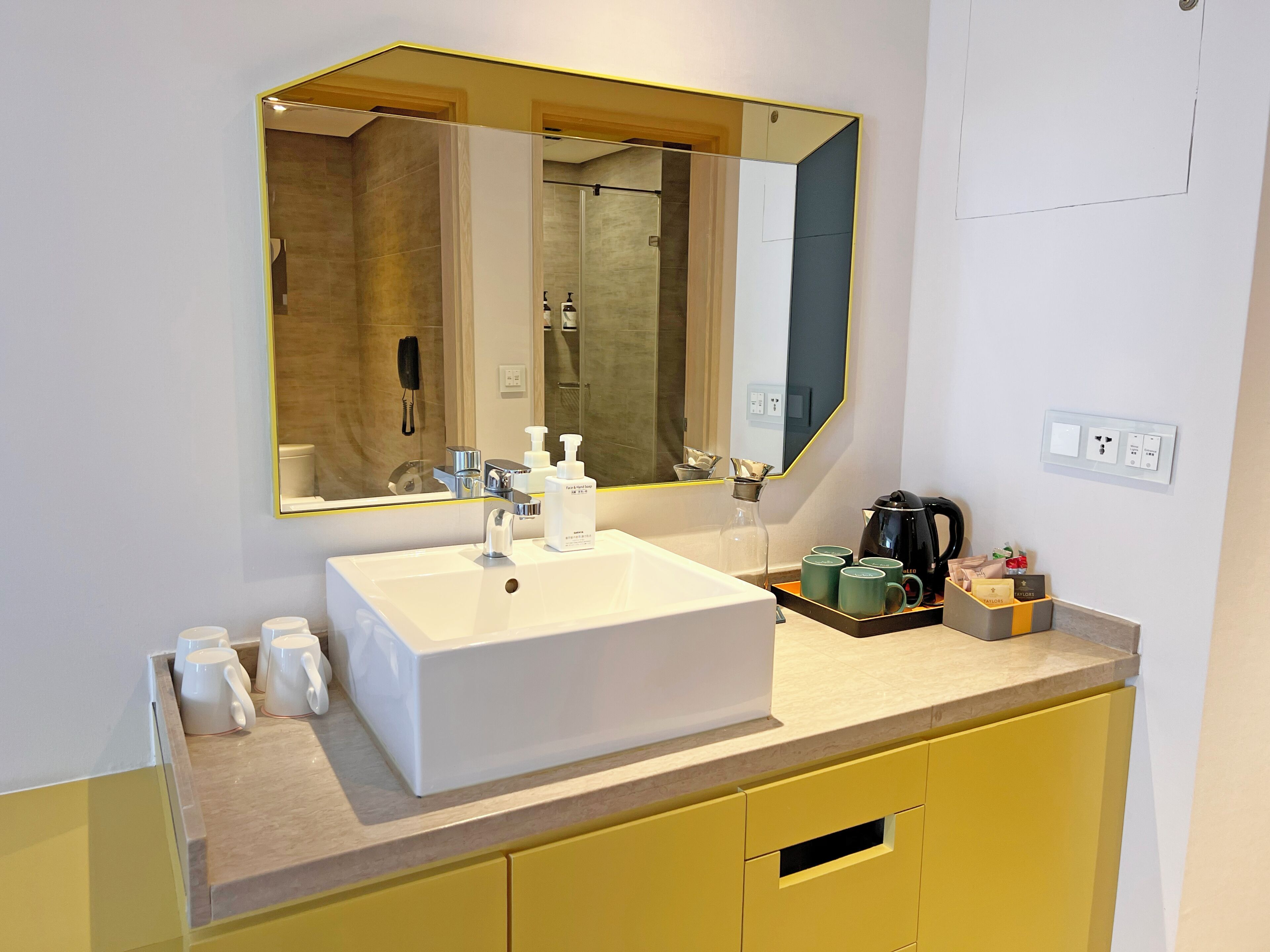 A modern bathroom with a large mirror, white sink, yellow cabinets, and a coffee maker on the counter.