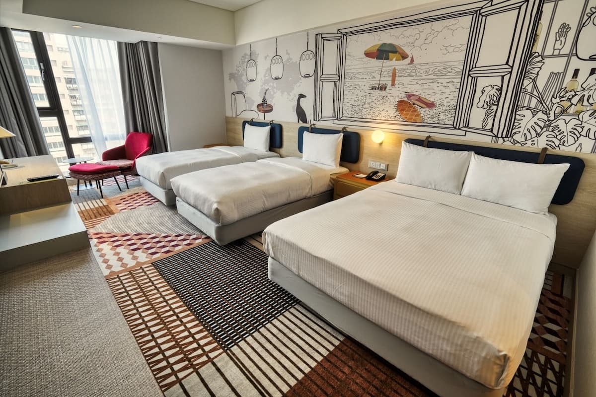 A hotel room with two beds, a red chair, a desk, and a large window.