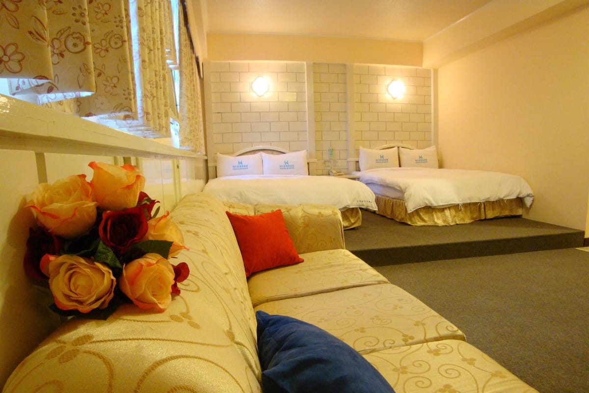 A hotel room with two beds, a sofa with a bouquet of roses, and a window with curtains.