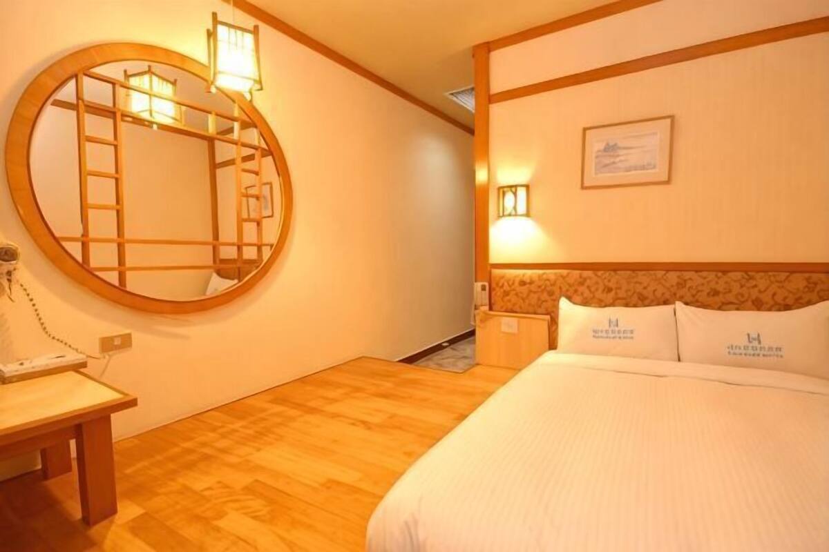 A hotel room with a wooden floor, a round mirror, a bed with white bedding, a nightstand, and a wall-mounted light fixture.