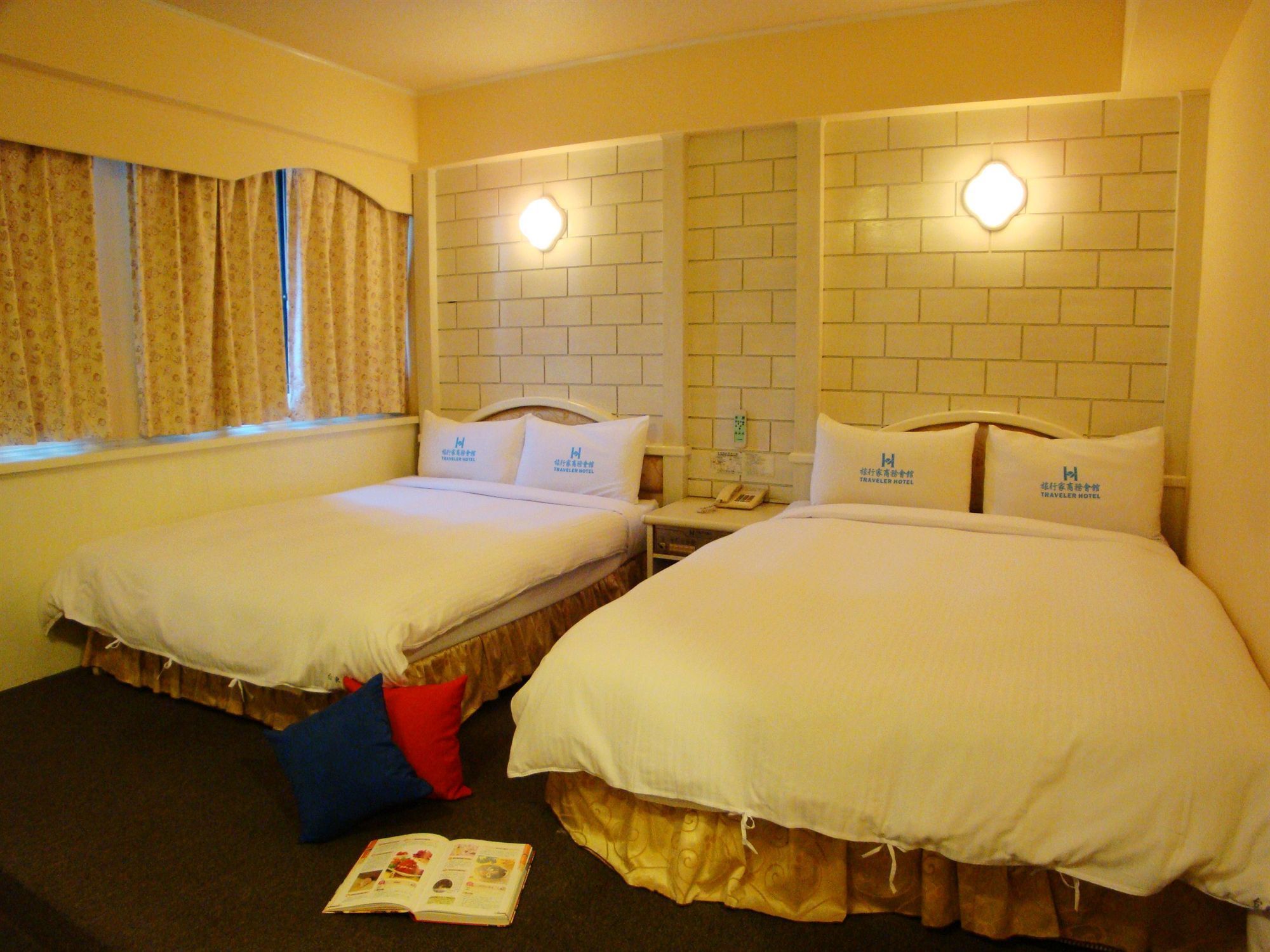 A hotel room with two beds, each with a pillow displaying the hotel logo, a bedside table with a phone, and a window with curtains.