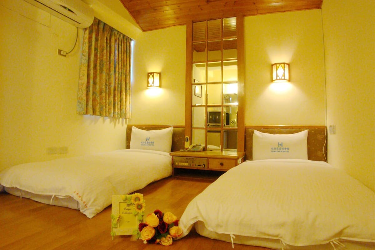 A hotel room with two beds, a wooden ceiling, a window with curtains, and wall-mounted lights.