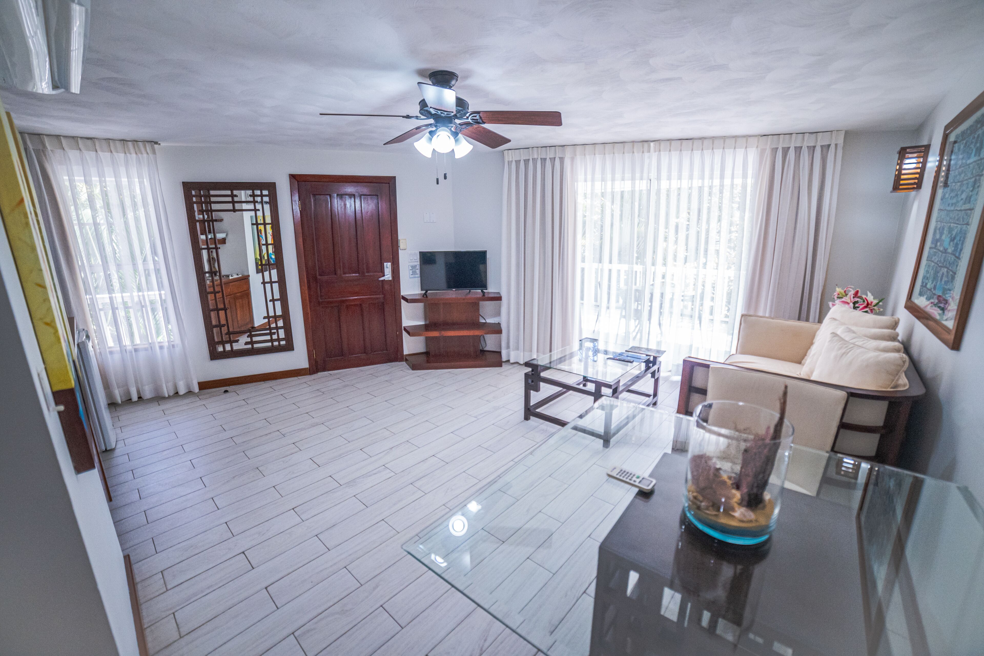 A spacious living room with a ceiling fan, a glass coffee table, a sofa, and a TV.