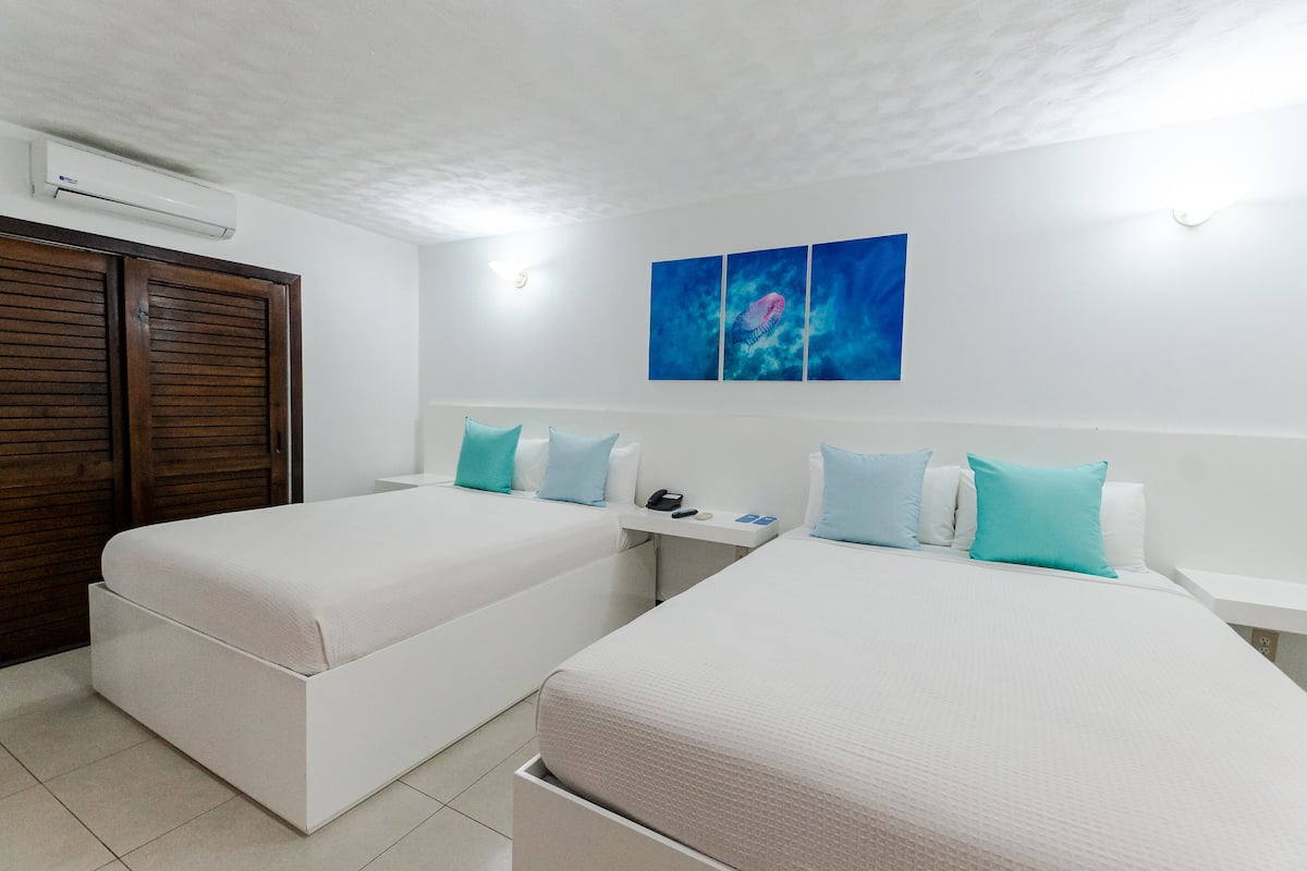 Habitación de hotel con dos camas individuales, un armario de madera, una mesita con un teléfono y una lámpara, y un aire acondicionado instalado en la pared.