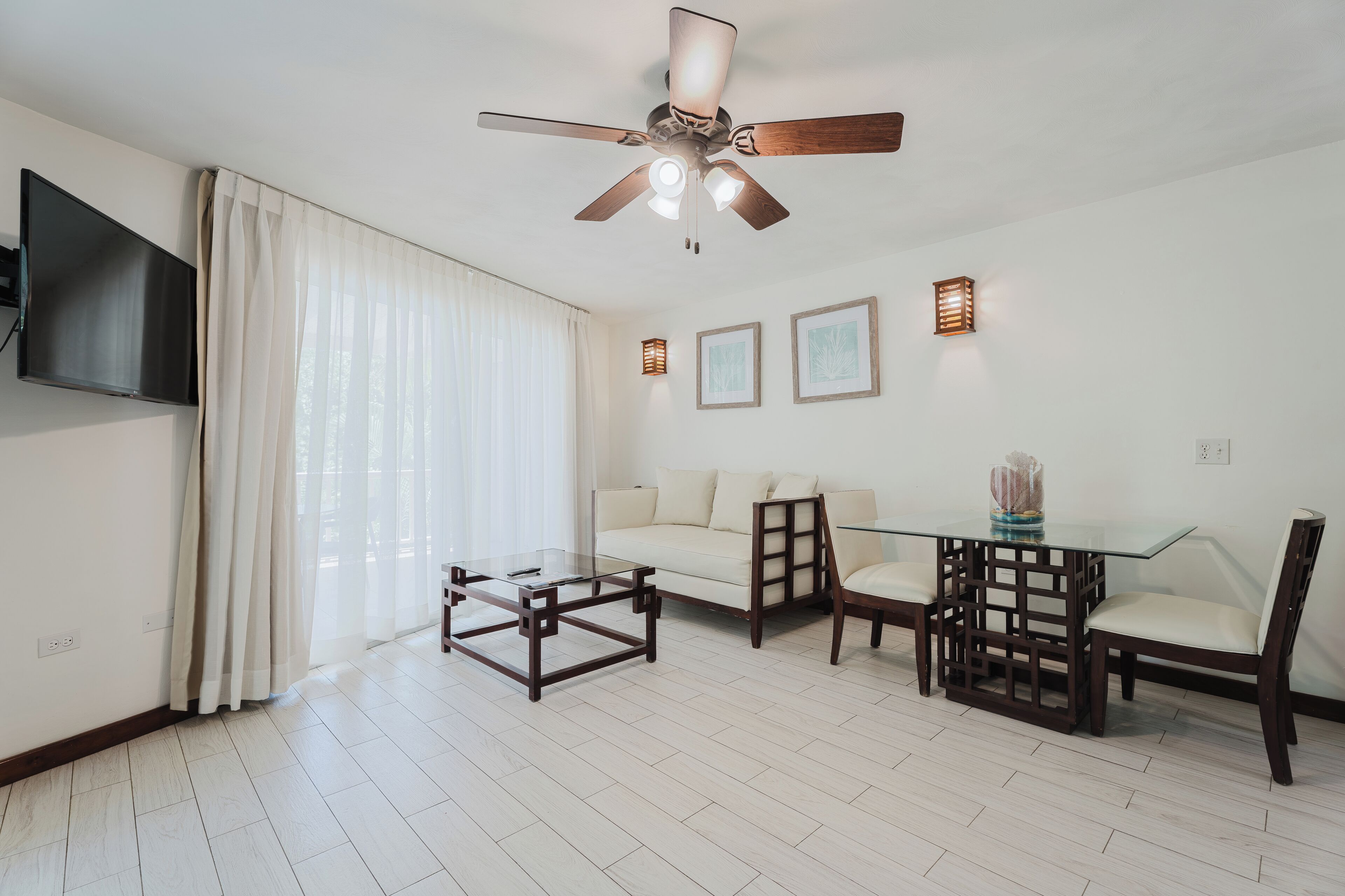 A modern living room with a ceiling fan, a flat-screen TV, a glass coffee table, a sofa, a dining table with chairs, and framed artwork on the wall.
