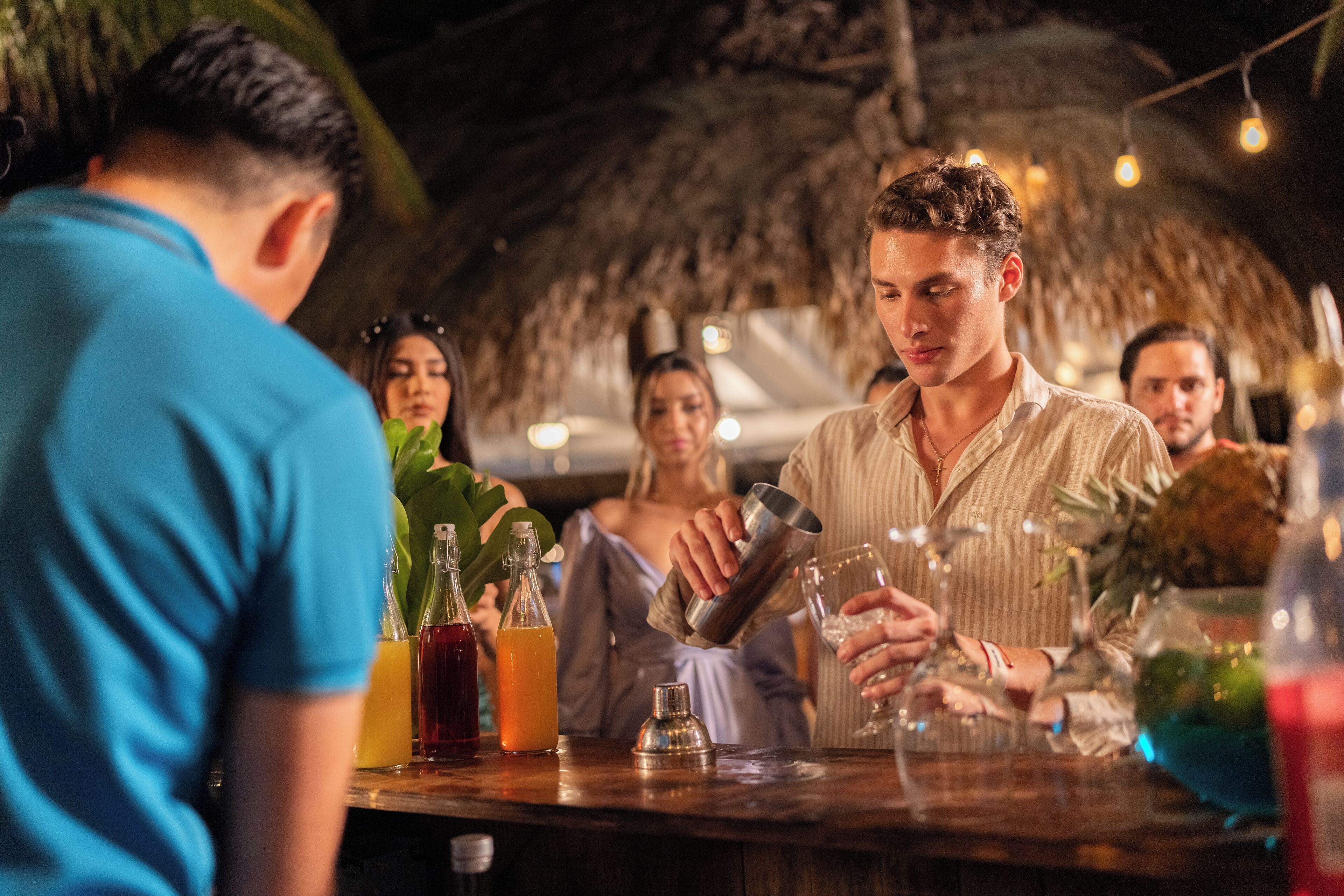 Eine Bar mit rustikalem Strohdach, einem Mixologen, der Getränke zubereitet, und verschiedenen Flaschen und Gläsern auf der Theke.