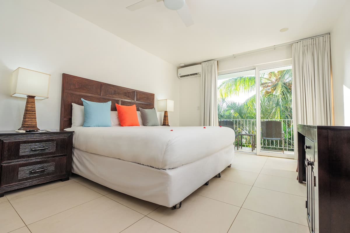 Habitación de hotel con una cama grande, cabecera de madera, mesita de noche con lámparas y un balcón con vistas a palmeras.