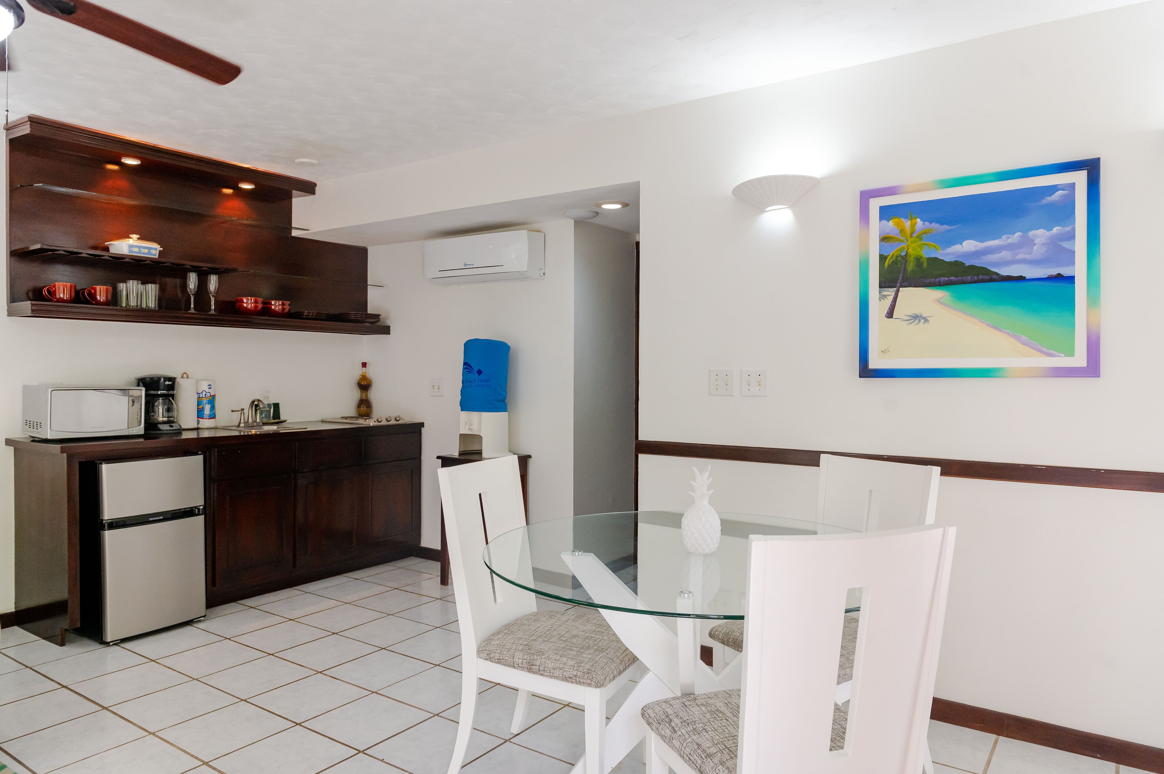 A modern kitchen with a built-in microwave, a refrigerator, and a coffee maker. There is a dining area with a glass table and white chairs.