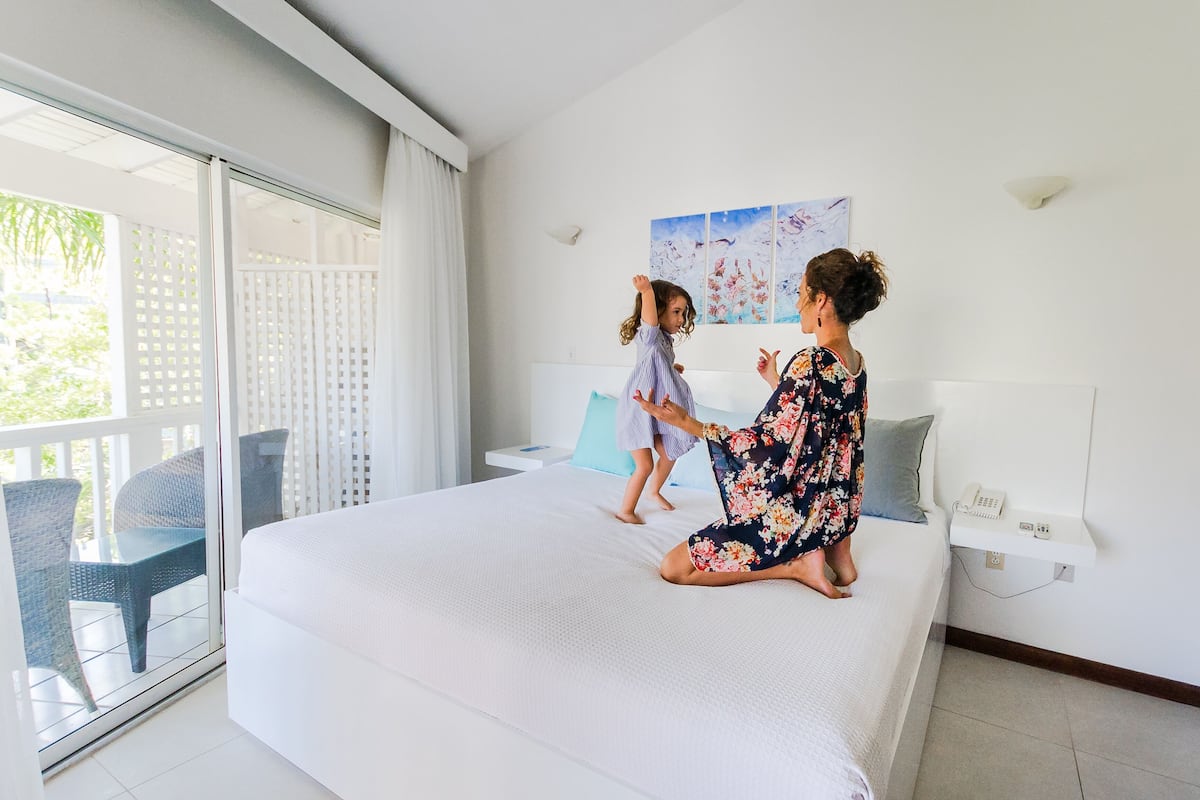Un dormitorio con cama, mesita de noche, teléfono en la pared y ventana con vista a follaje.