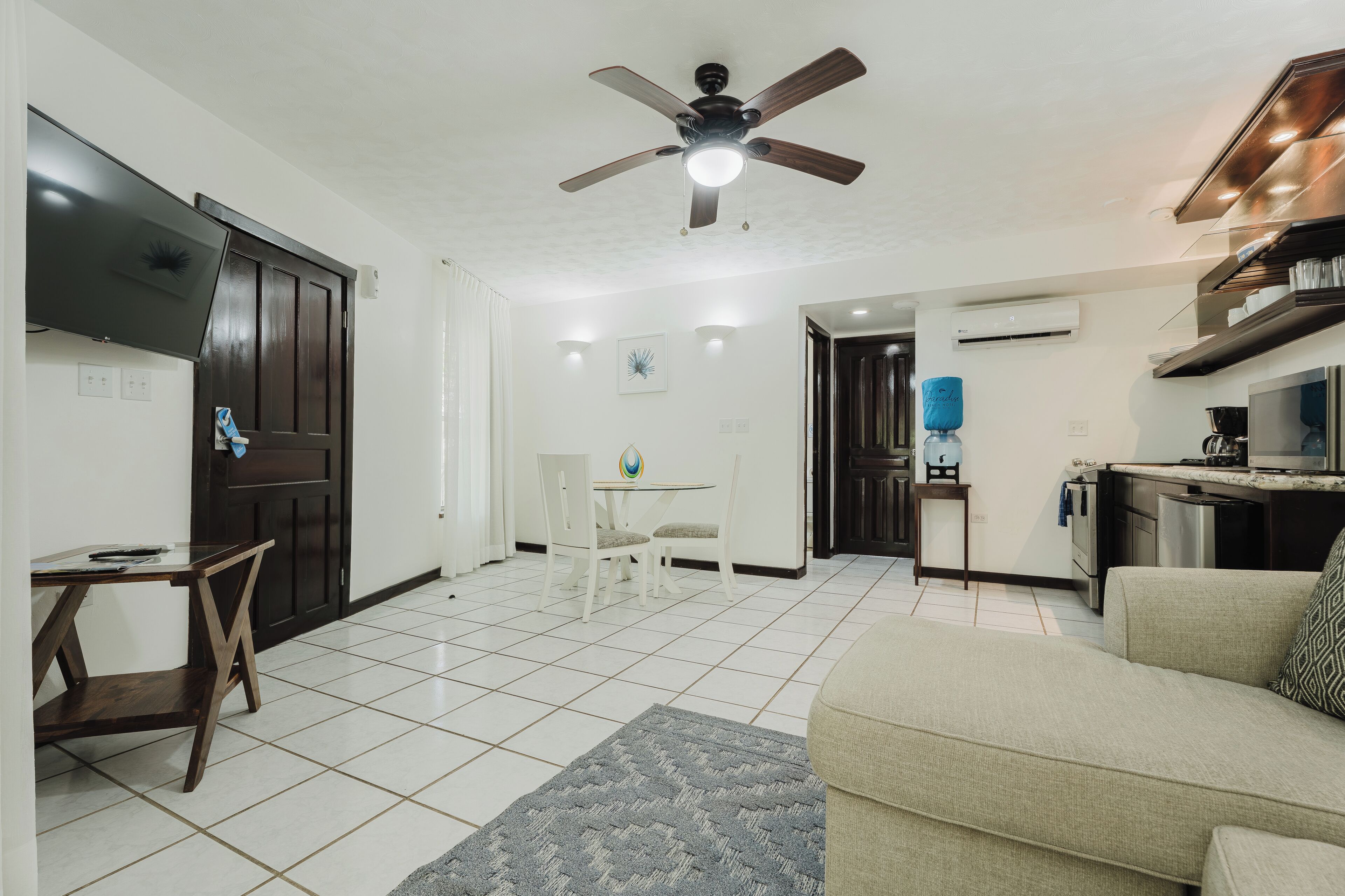 A living room with a sofa, a coffee table, and a TV. There is a dining area with a table and chairs. The kitchen has a stove and a water dispenser.