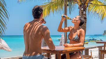 Una pareja sentada en una mesa de madera en la playa, con vista despejada al océano y barcos al fondo.