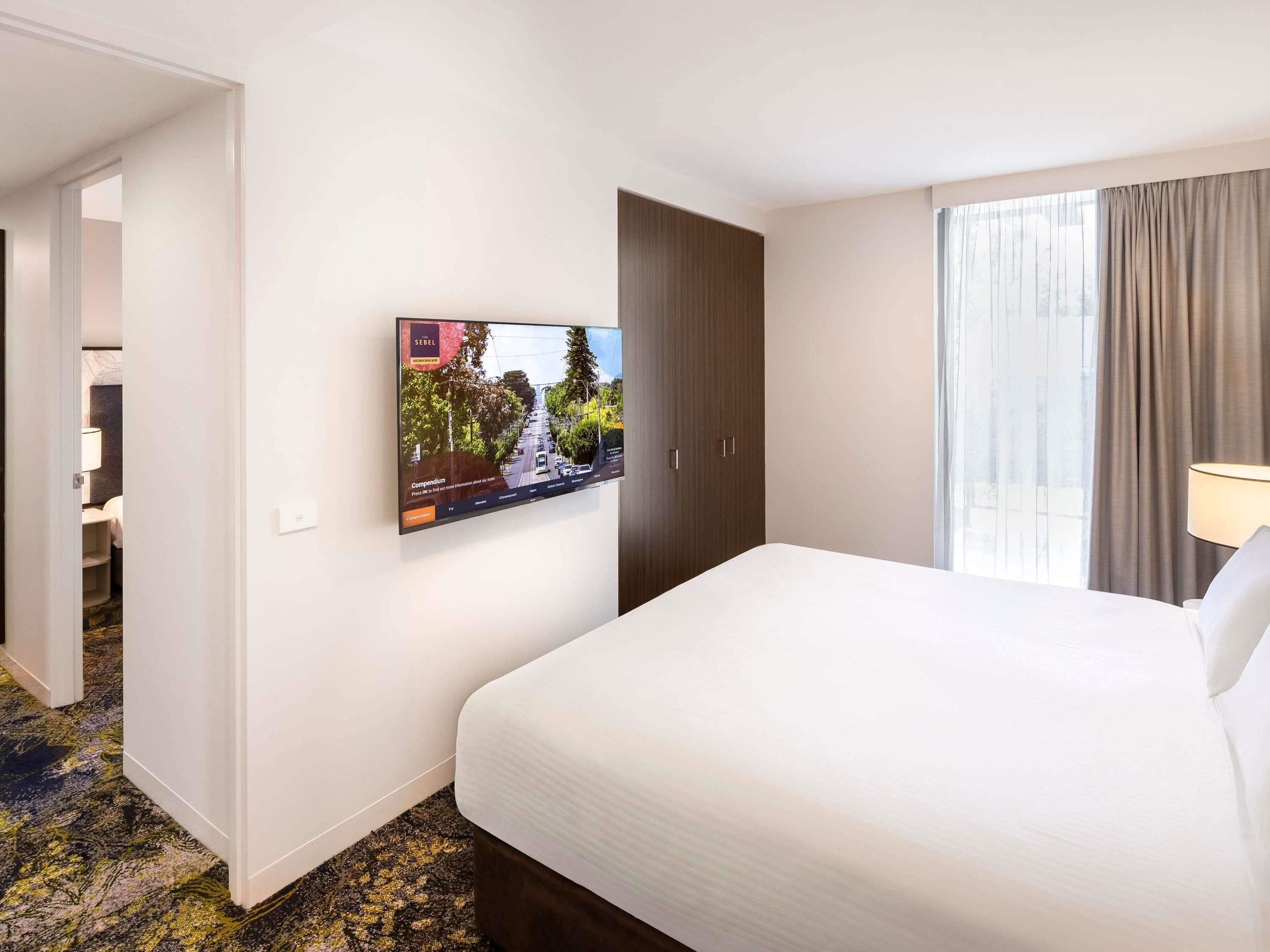 Habitación de hotel con cama, televisor montado en la pared, armario y ventana con cortinas.