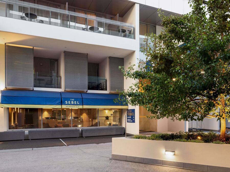 Un hotel moderno con un toldo azul, balcones de vidrio y un árbol en primer plano.