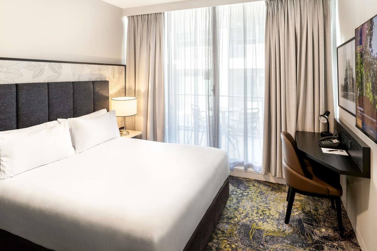 Una habitación de hotel con una cama, un escritorio, una silla, una lámpara y un ventanal con cortinas.