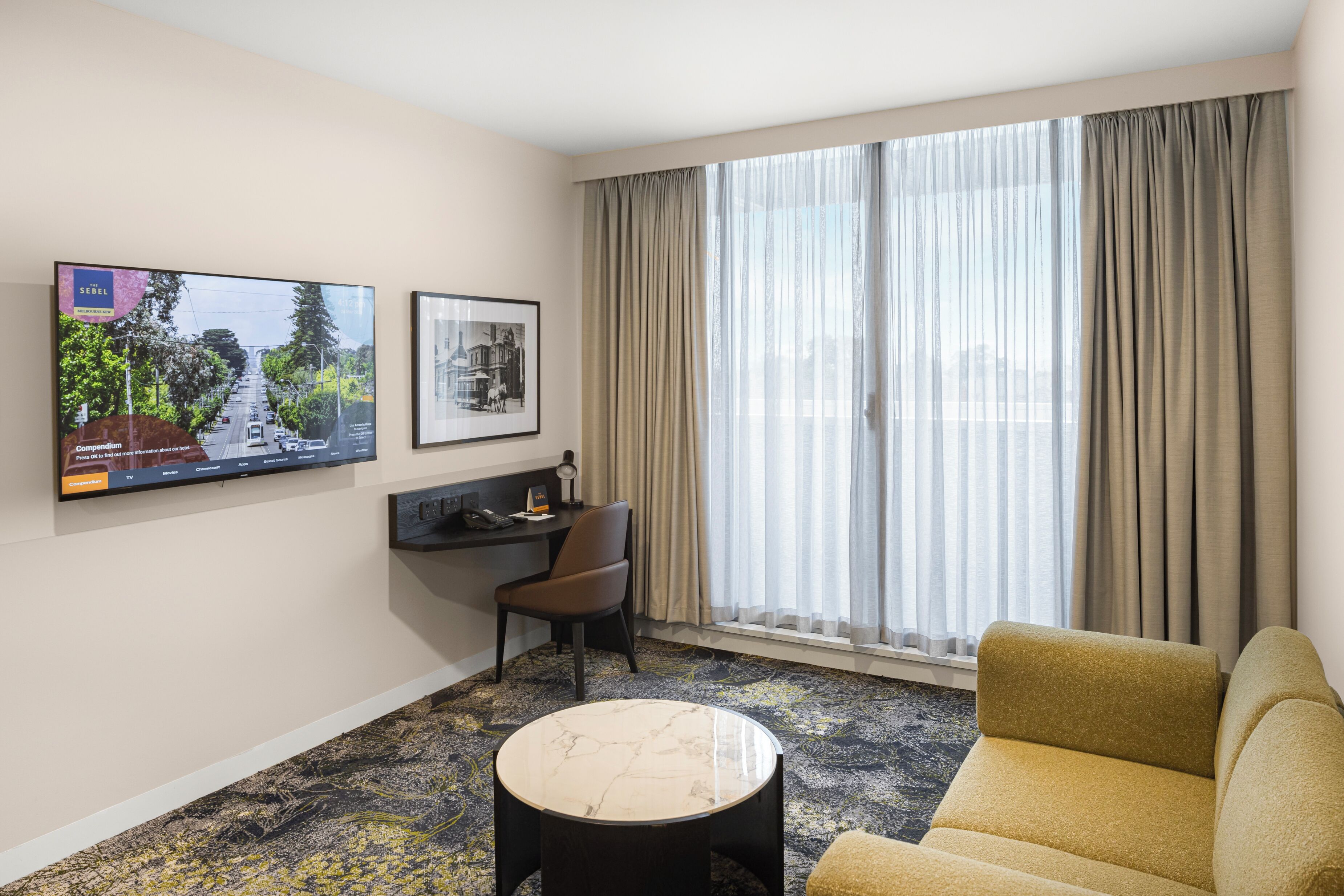 Een hotelkamer met een bank, een rond bijzettafeltje, een bureau met stoel, een aan de muur bevestigde televisie en een groot raam met gordijnen.