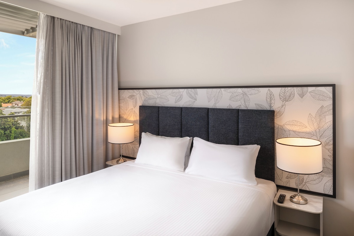 A hotel room with a bed, two bedside lamps, a nightstand, a remote control, and a view of greenery outside.