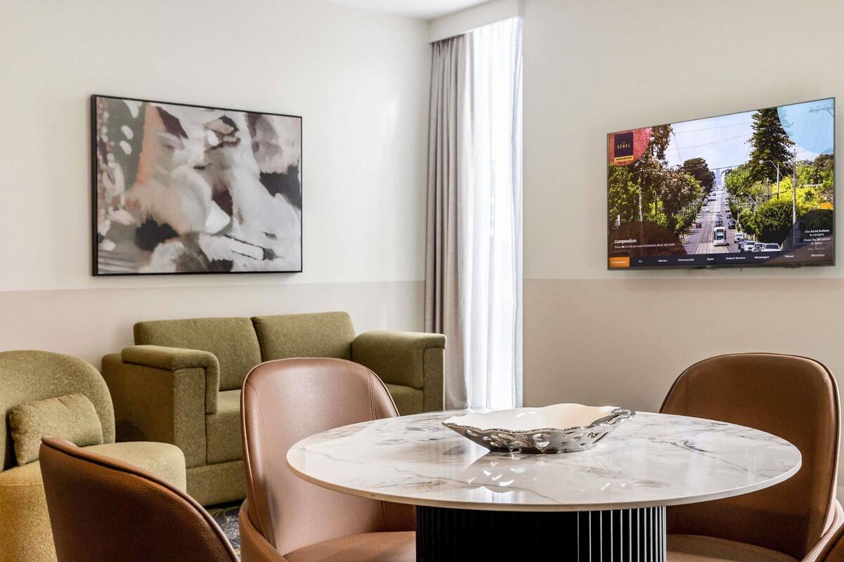 Quarto de hotel moderno com uma mesa redonda de mármore, poltronas de couro, um sofá verde, uma grande pintura abstrata e uma televisão exibindo uma vista urbana.