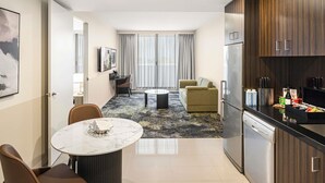A modern hotel room with a dining area, a sofa, a kitchen with a refrigerator and utensils, and a large window with curtains. - The Sebel Melbourne Kew (Kew)