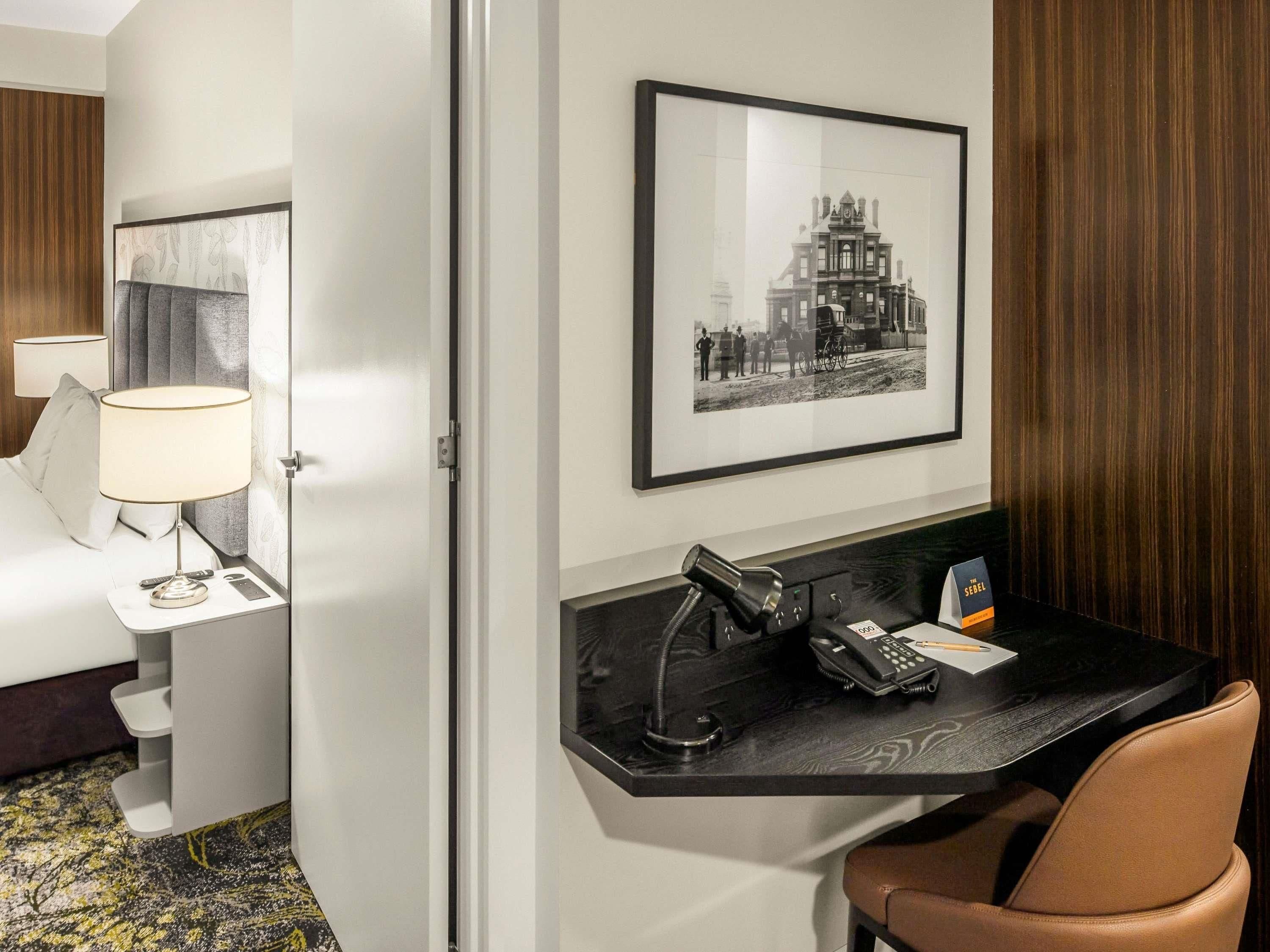 Habitación de hotel con cama, mesita de noche con lámpara, escritorio con teléfono y un cuadro en la pared.