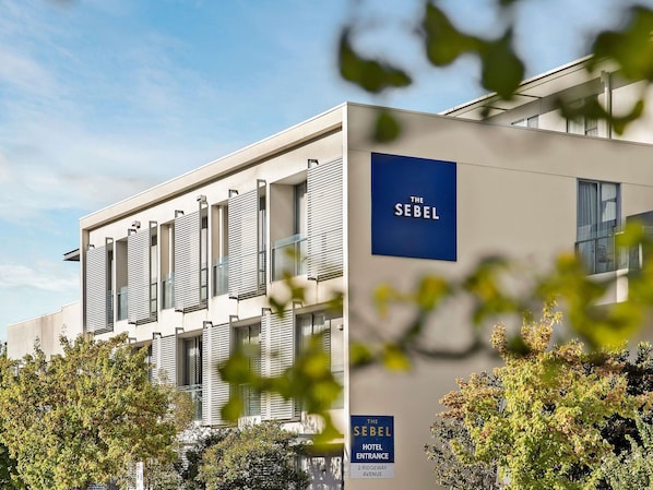 A modern hotel building with a sign reading 'The Sebel Hotel Entrance' and '2 Ridgeway Avenue'.