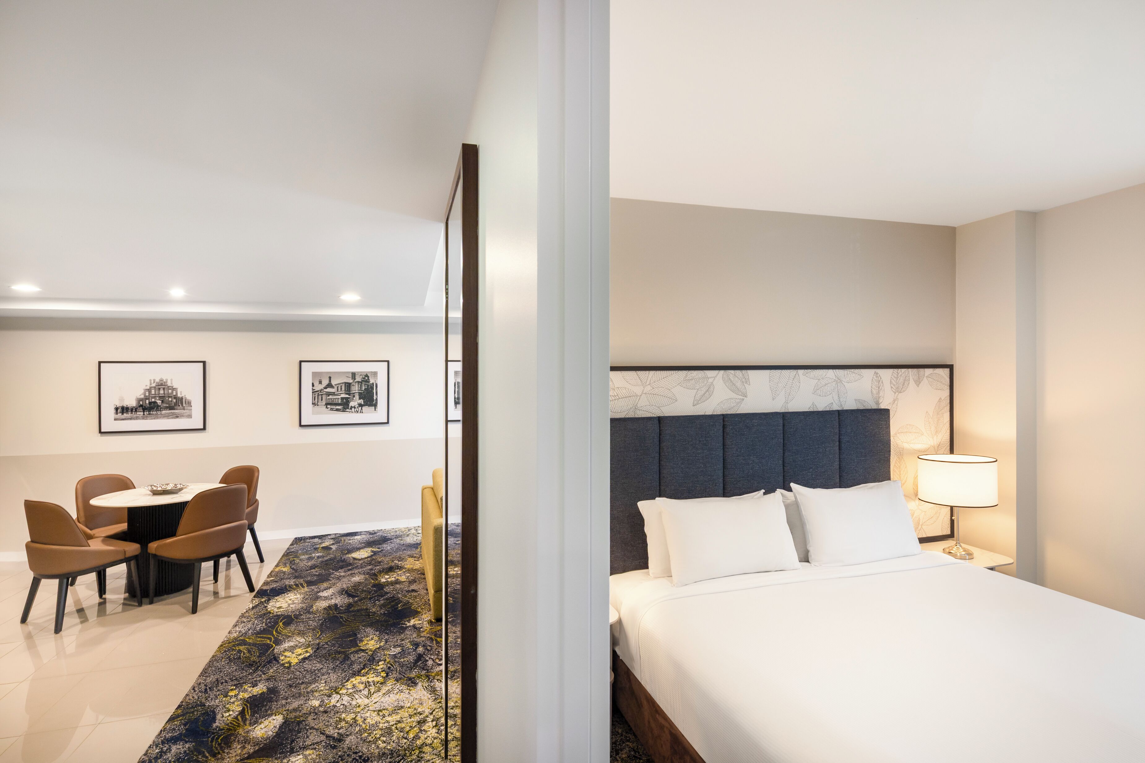 A hotel room with a bed, a bedside table with a lamp, a dining area with chairs, and framed pictures on the wall.