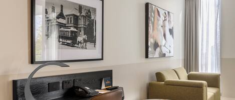 A hotel room with a desk, chair, sofa, artwork, and a window with curtains.