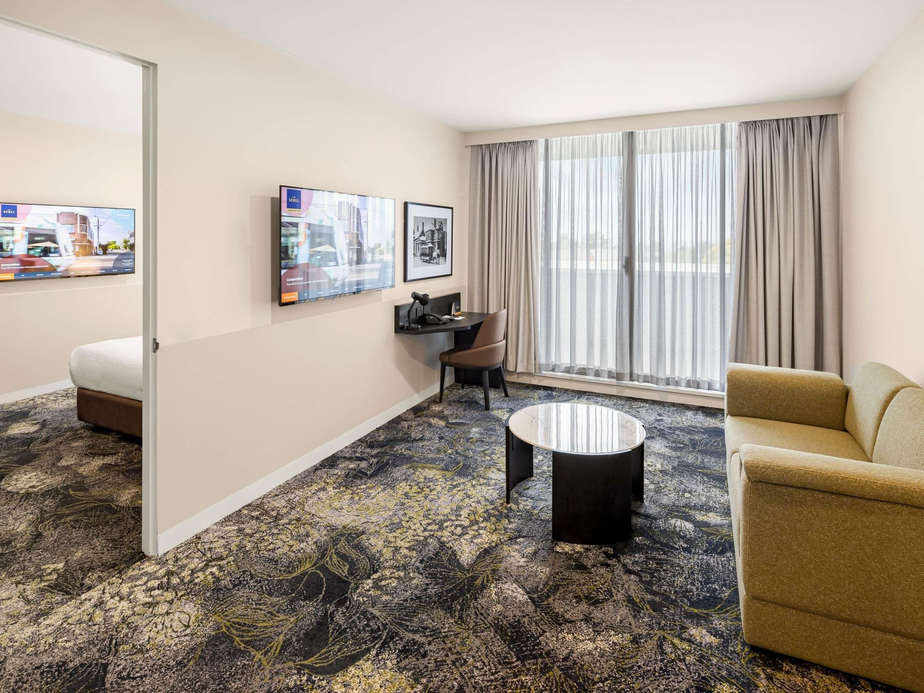 A hotel room with a bed, a desk, a chair, a sofa, a coffee table, and a large window with curtains.