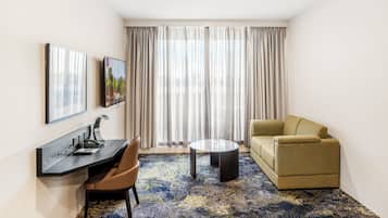 A hotel room with a desk, a sofa, a coffee table, and a large window with curtains.
