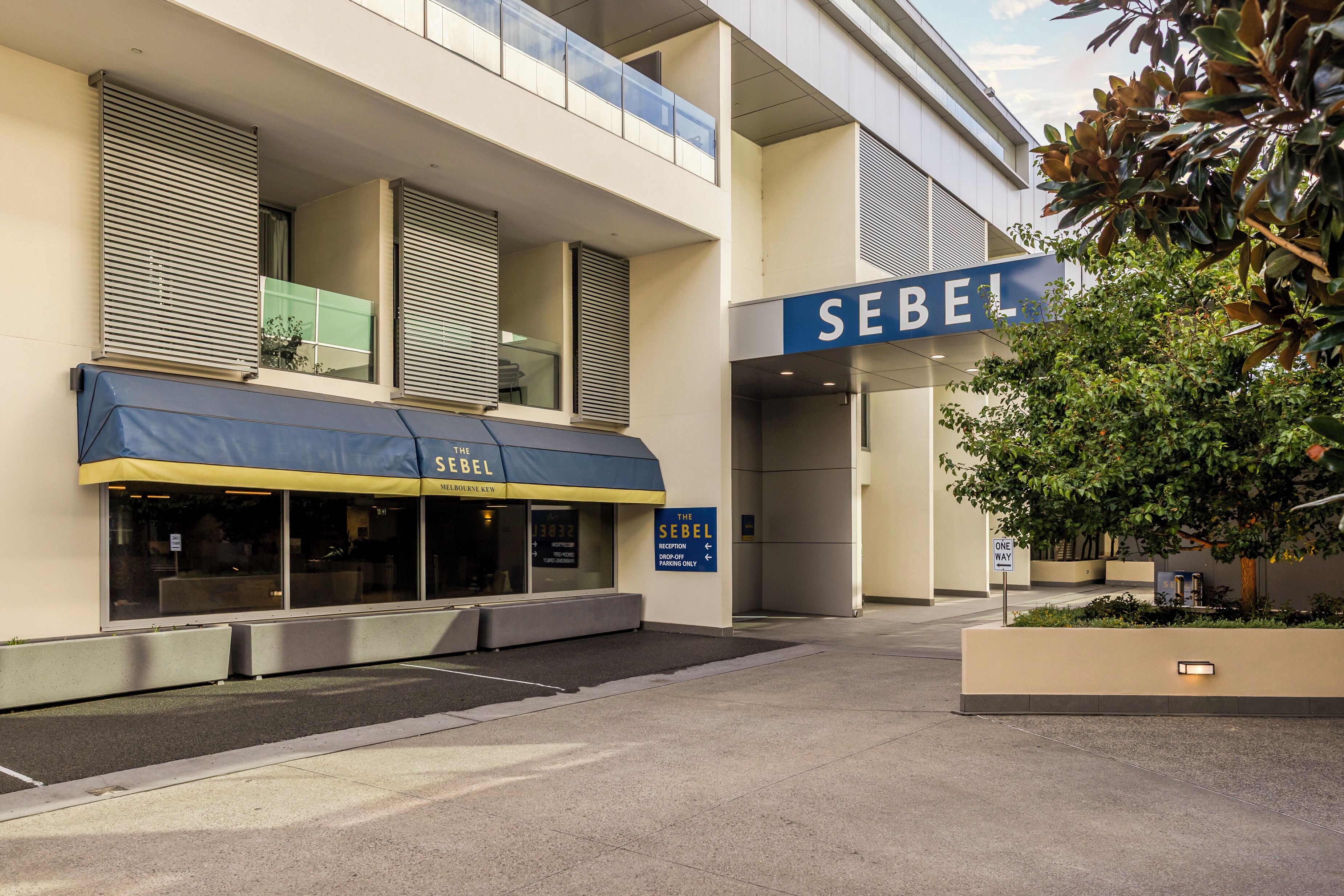 A modern building with a 'SEBEL' sign, a covered outdoor seating area, and a 'ONE WAY' parking sign.