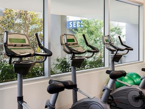 Three stationary bikes with digital displays, a green exercise ball, and a window with a view of trees and a sign reading 'SEBA'. - The Sebel Melbourne Kew (Kew)