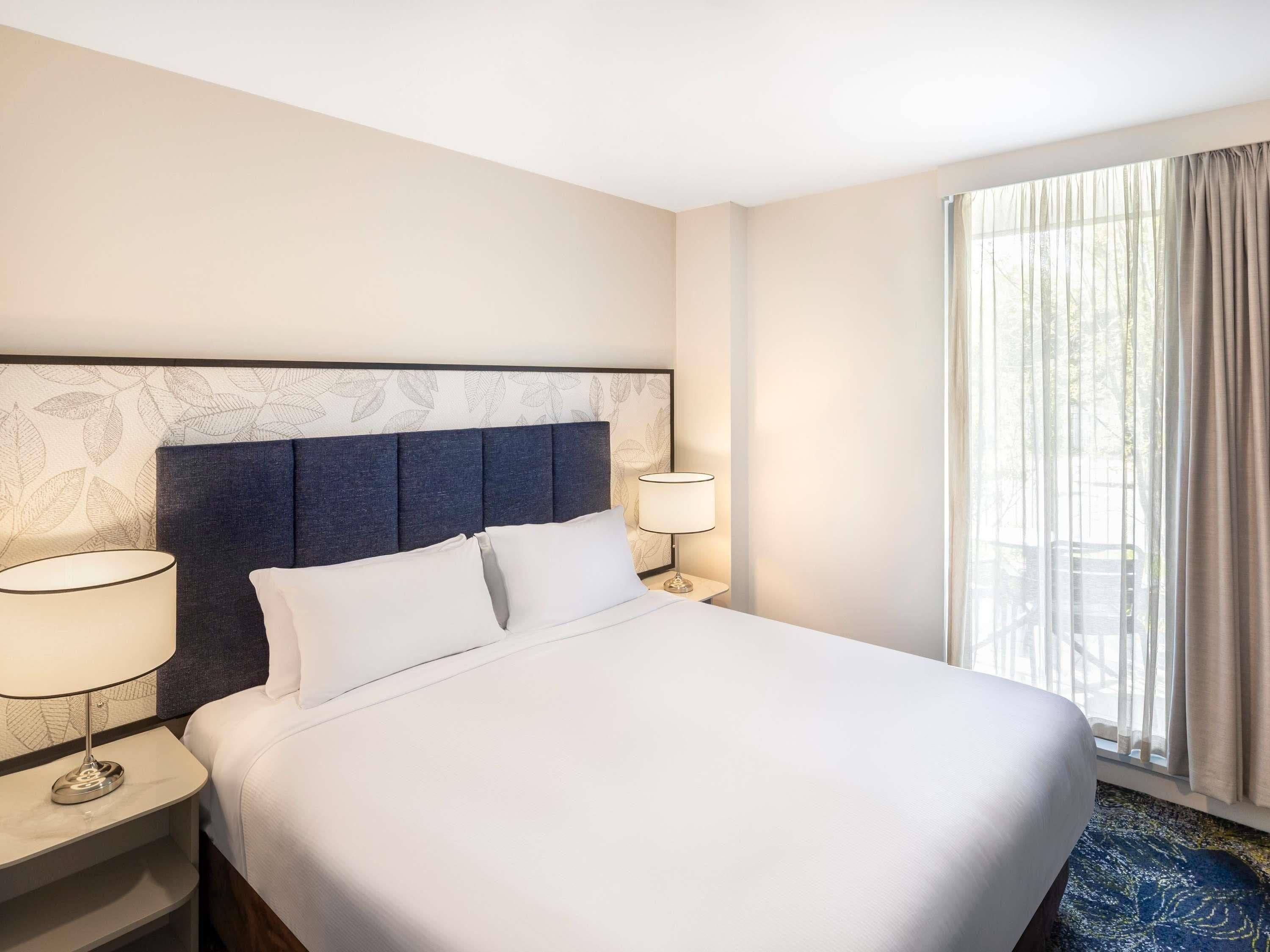 A hotel room with a large bed, two bedside tables, a nightstand, a lamp, and a window with curtains.