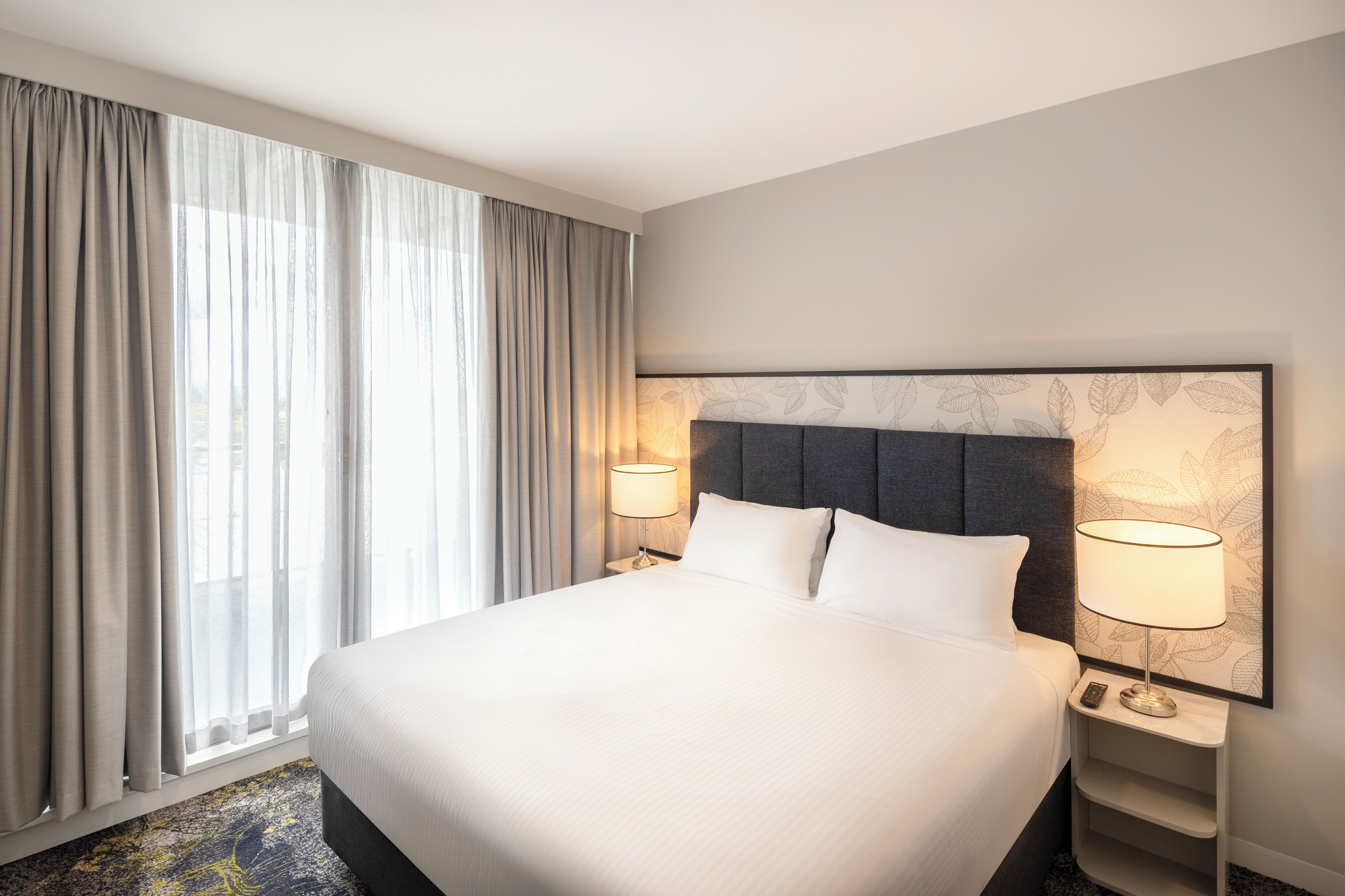 A hotel room with a large bed, two bedside tables with lamps, a window with curtains, and a wall with a leaf pattern.
