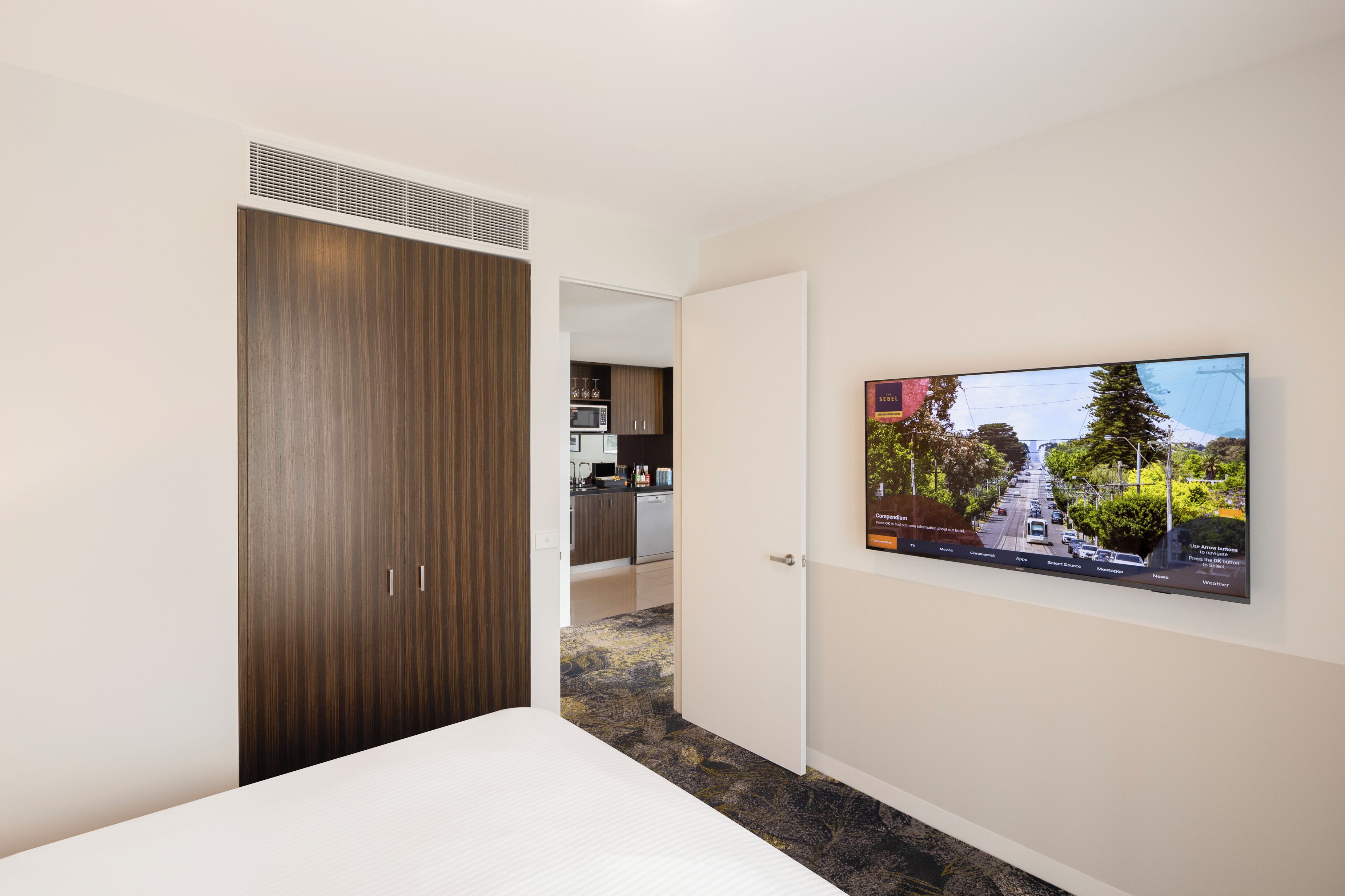 A hotel room with a bed, a wardrobe, a television mounted on the wall, and a view of the kitchen area.