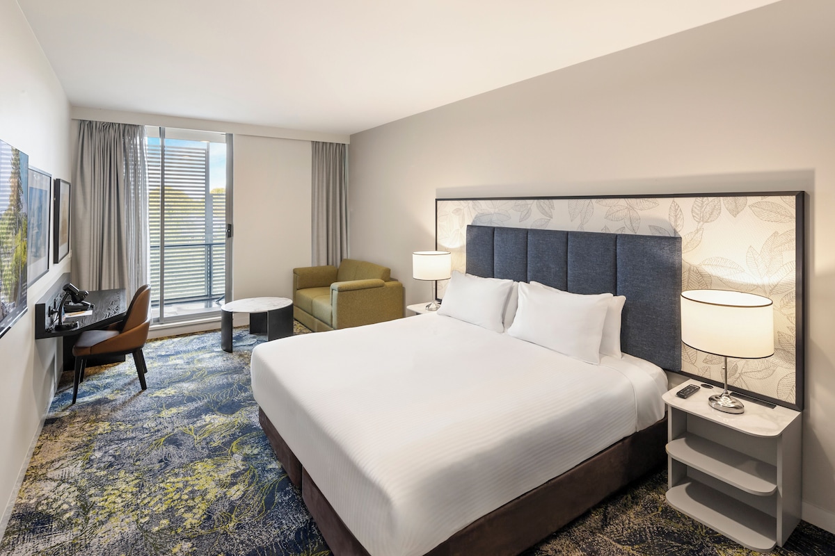 A hotel room with a large bed, a desk, a chair, a small table, and a nightstand with a lamp.