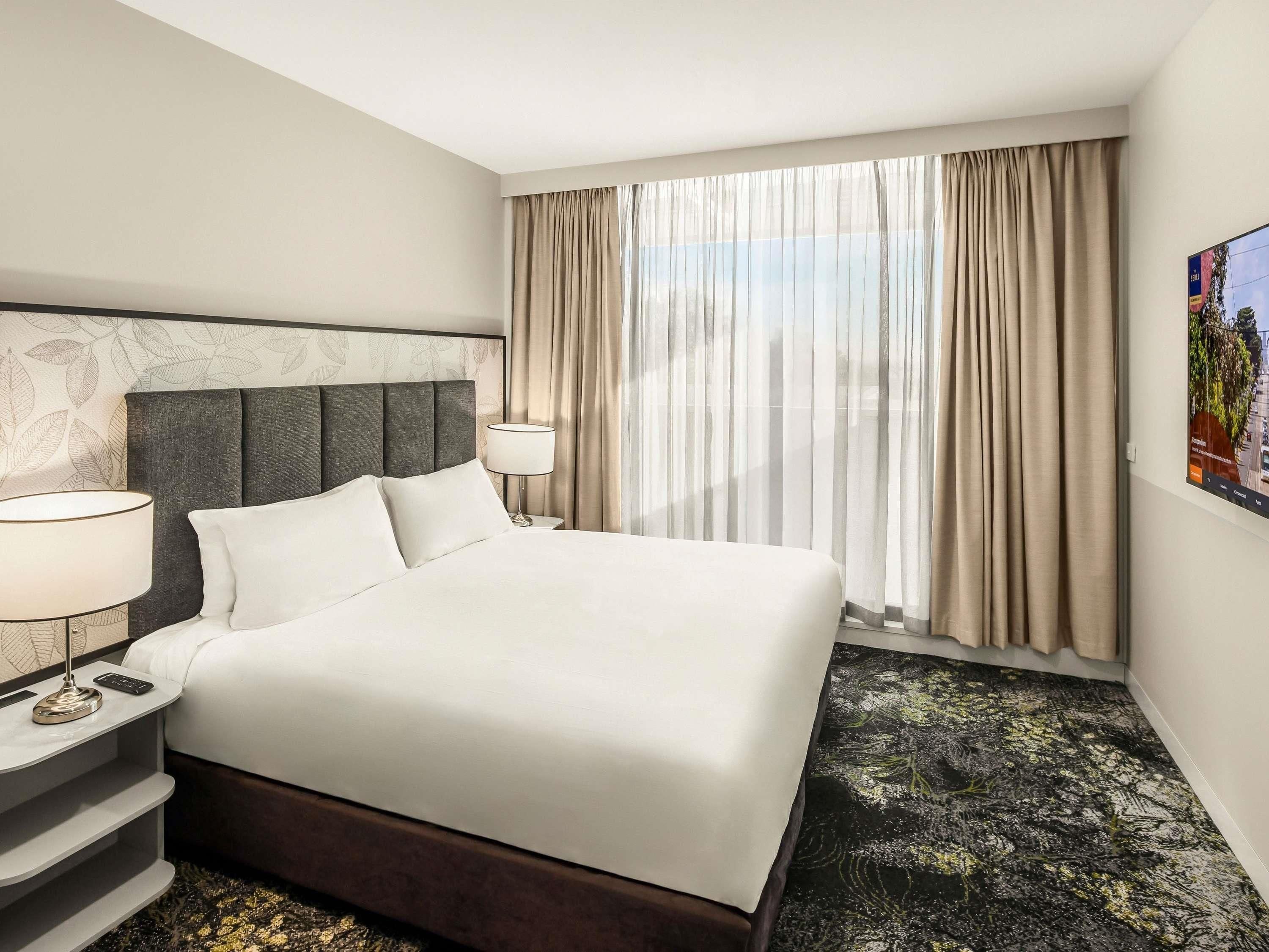 A hotel room with a large bed, a nightstand with a lamp, a headboard, a window with curtains, and a wall-mounted painting.