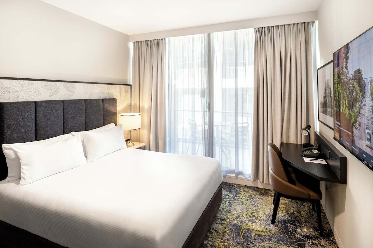 A hotel room with a bed, a desk, a chair, a lamp, a painting, and a view of the city.