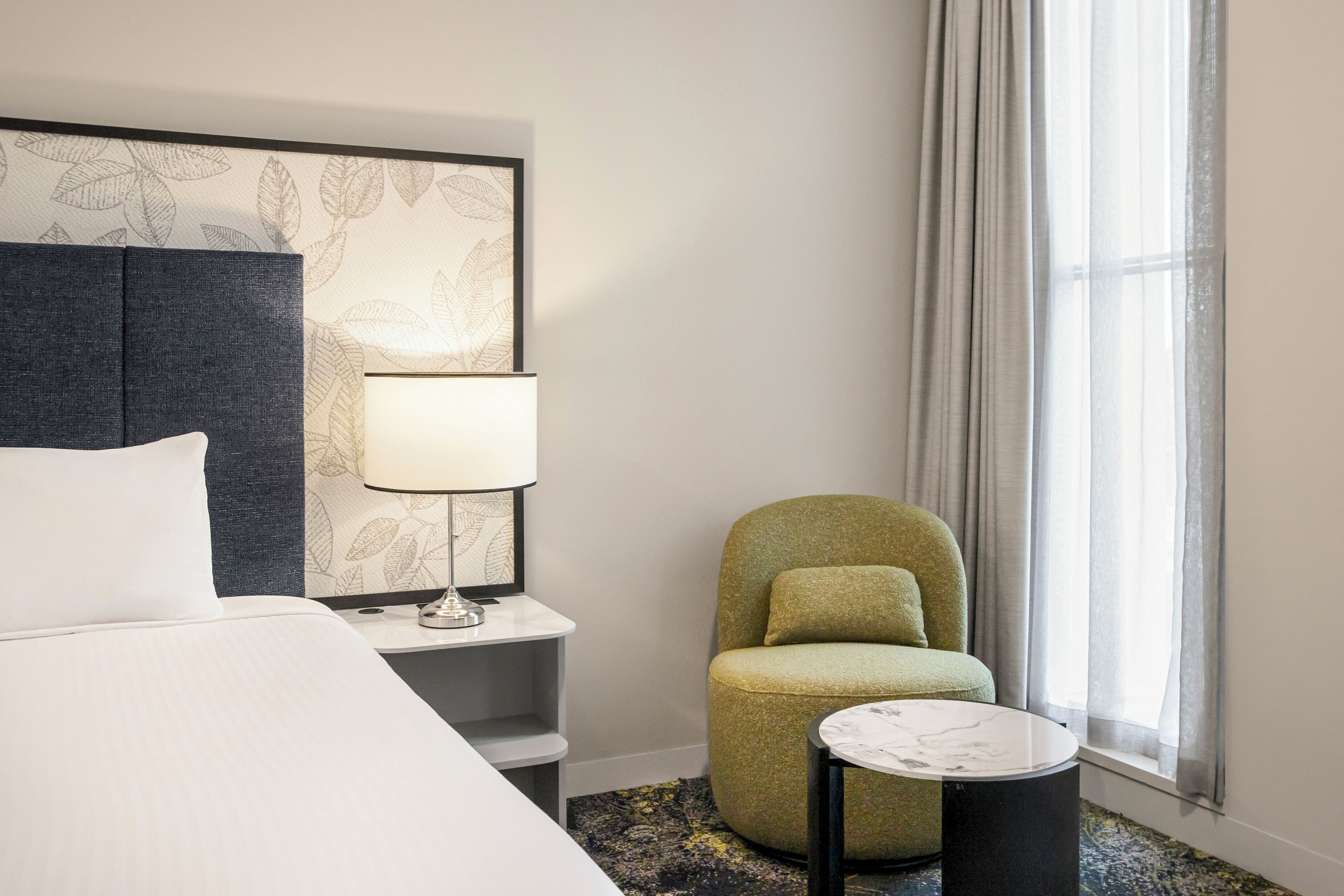 A hotel room with a bed, a nightstand with a lamp, a green chair, a small round table, and a window with curtains.
