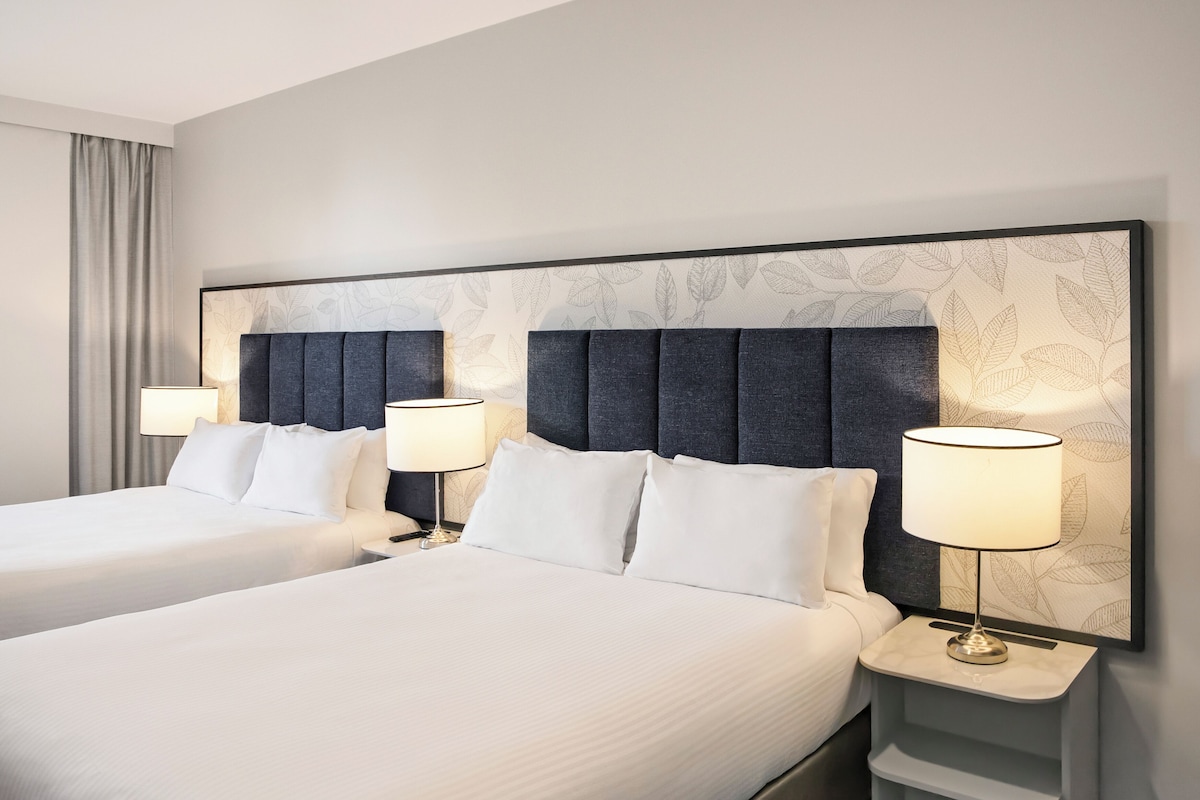 A hotel room with two beds, a large headboard, bedside lamps, and a wall with a leaf pattern.