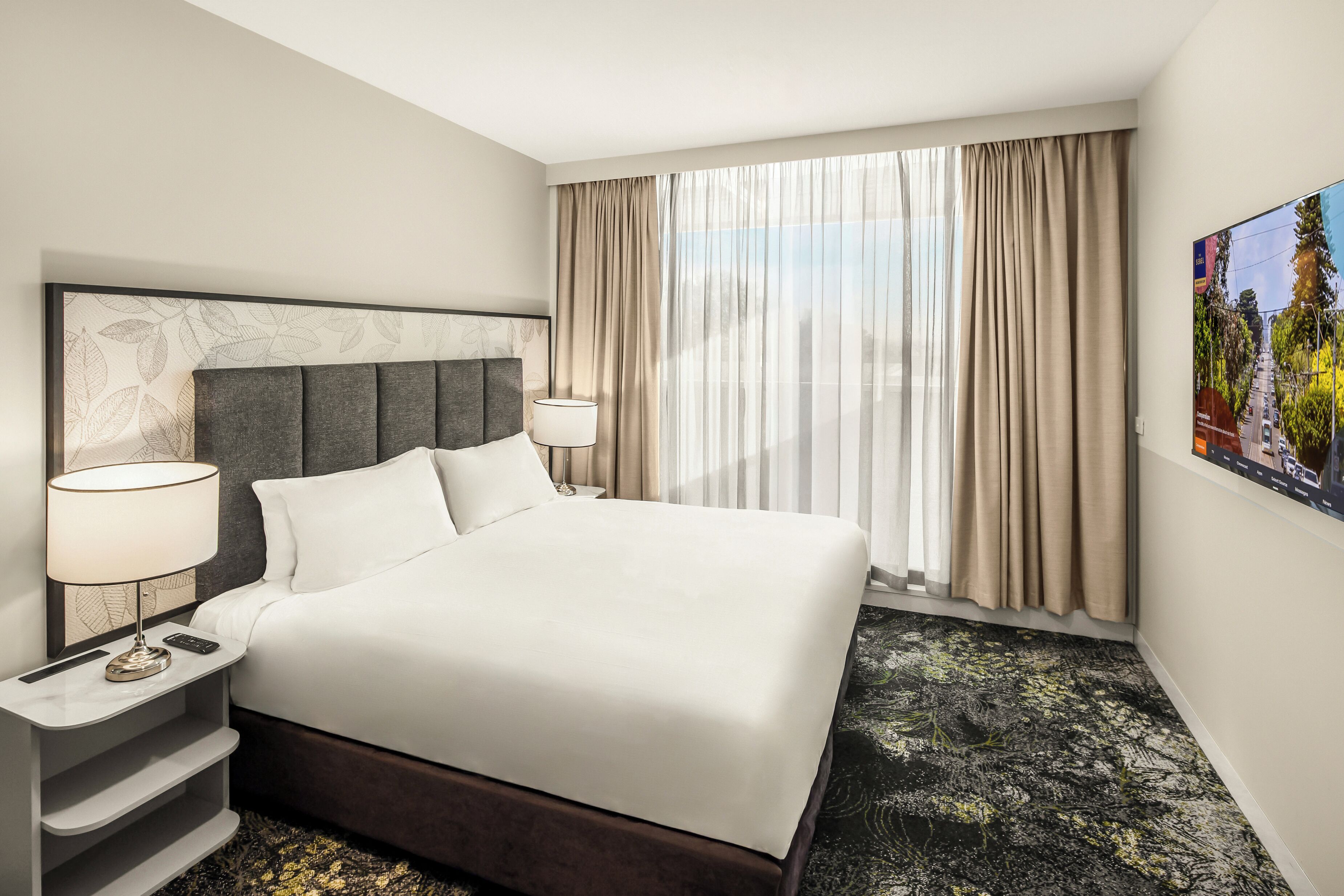 A hotel room with a large bed, a headboard with leaf patterns, a nightstand with a lamp, and a large window with curtains.