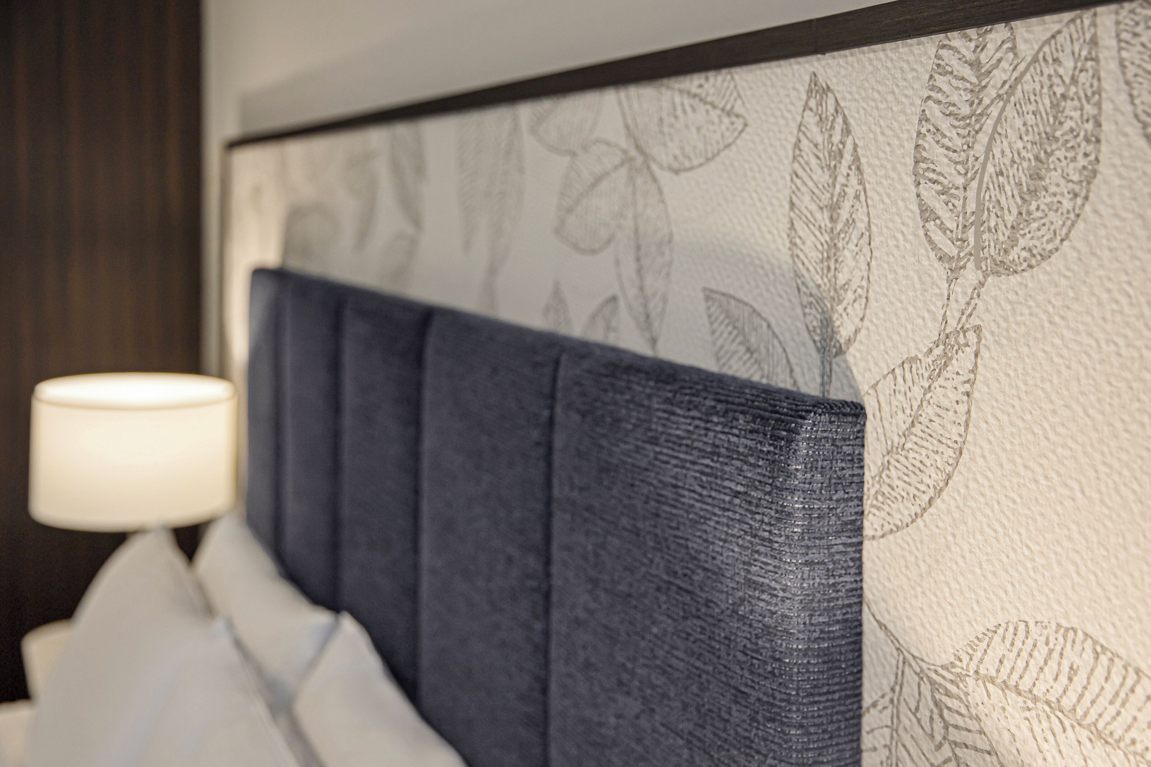 A headboard with a leaf pattern, a white lamp, and a dark-colored wall.
