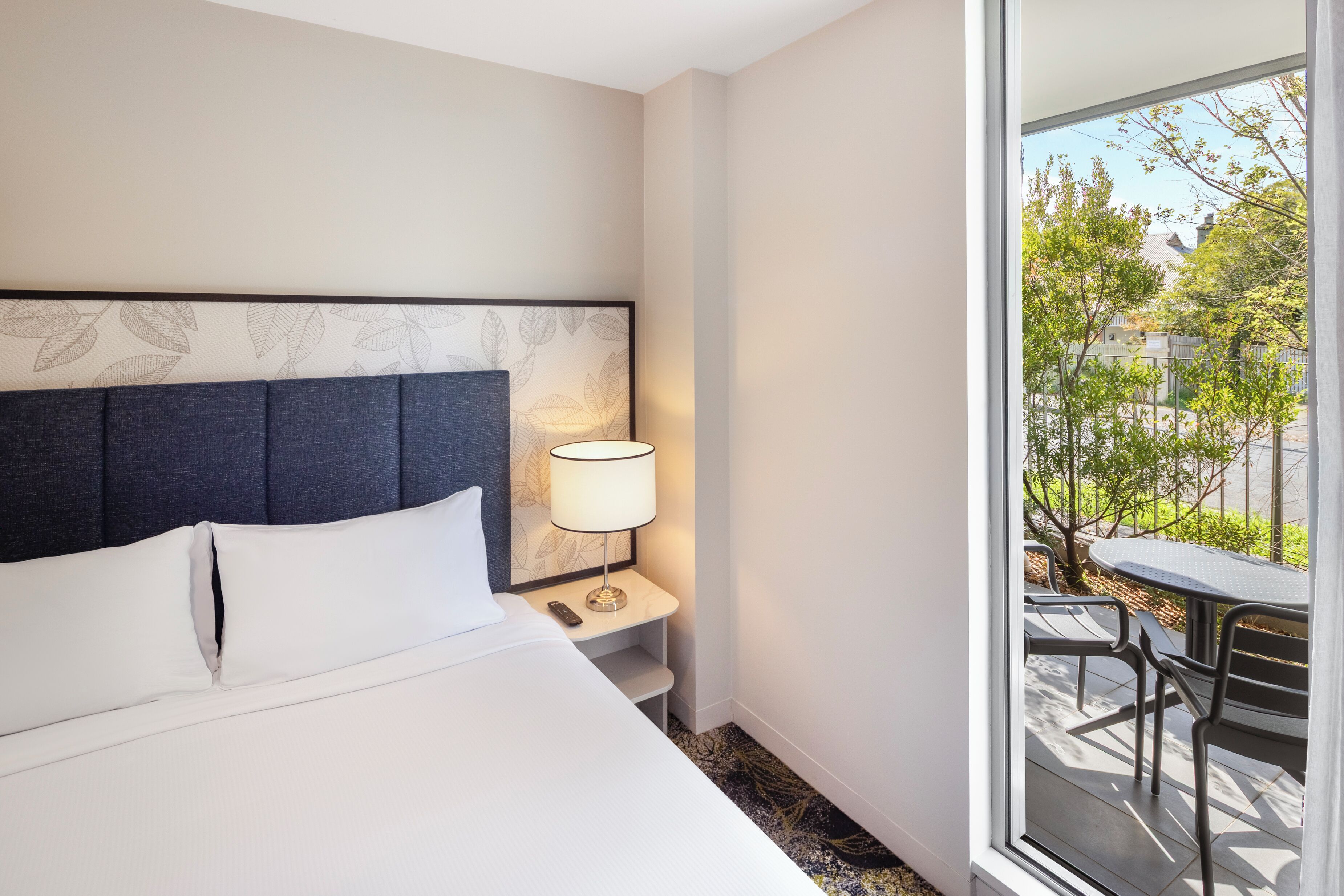 A hotel room with a bed, a nightstand with a lamp, and a view of the outdoors through a large window.